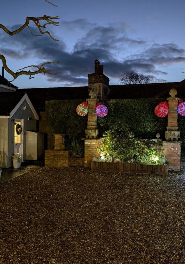 The Cottage at Christmas with lights, reindeer, a tree, wreath, and large red ornaments.